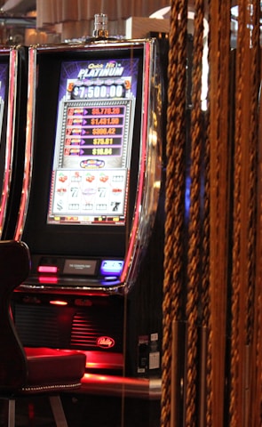 A pair of casino slot machines, each with detailed screens displaying various game outcomes and jackpot amounts. The chairs in front of the slot machines are dark and cushioned for comfort. The background features a hanging curtain made of golden ropes and other casino decorations are visible.