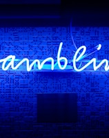 A bright blue neon sign spells out the word 'gambling' in cursive script. The background features a textured wall covered in domino tiles, also illuminated by the blue light, creating a vivid and visually striking scene.