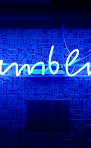 A bright blue neon sign spells out the word 'gambling' in cursive script. The background features a textured wall covered in domino tiles, also illuminated by the blue light, creating a vivid and visually striking scene.