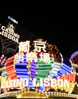 Bright neon signs in multiple colors illuminate the night, prominently displaying the name 'Casino Lisboa' alongside signage in Chinese characters. The lights form intricate patterns and vibrant displays, creating a lively and energetic atmosphere.