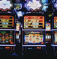 A row of colorful slot machines lit with bright neon lights, showcasing various graphical elements on the screens, indicating different games and jackpots. The atmosphere suggests a lively casino setting, bustling with excitement.