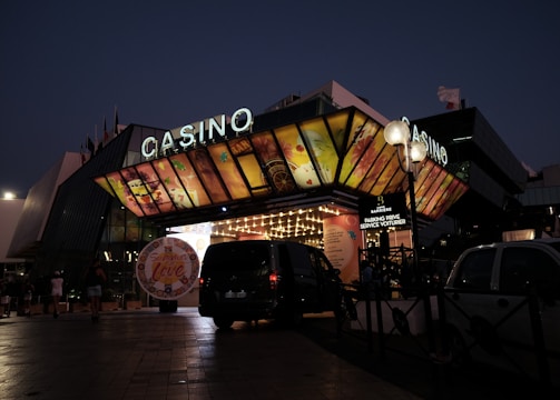 A building with a brightly illuminated entrance is seen at night. The sign above reads 'Casino' in bright letters. The facade features colorful graphics depicting various indulgent themes. Several vehicles are parked near the entrance, and people are visible in the vicinity. Tall lamp posts with spherical lights are present, casting a warm glow.