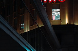 A night scene featuring a building with illuminated neon signage displaying 'CASINO'. The environment is urban with reflections on glass windows and glowing street traffic lights below. The atmosphere is dimly lit, with artificial lighting casting vibrant colors.
