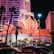 A brightly lit casino with neon lights in vibrant red, pink, and blue, featuring a large flamingo signage. Palm trees are visible in the foreground with vehicles passing by on the street at night.